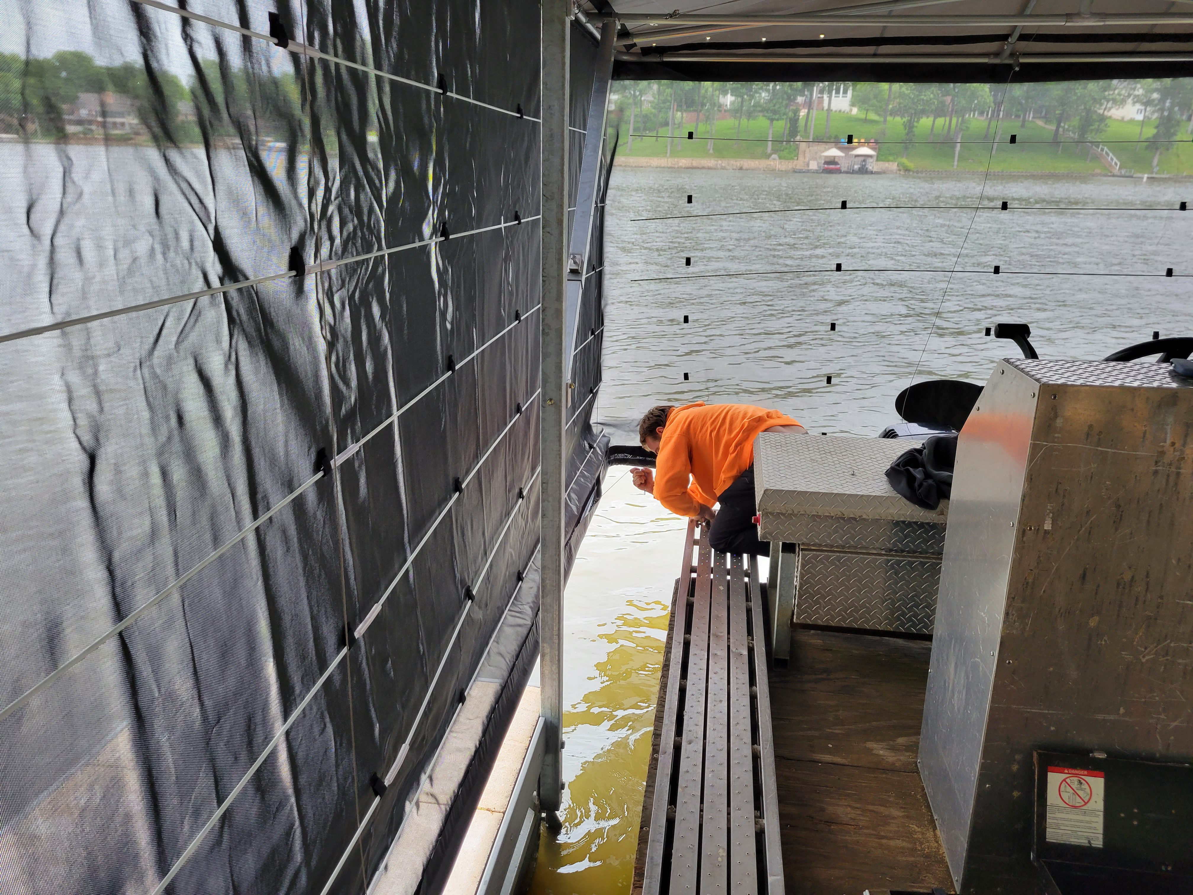 Person inspecting the side of a covered boat on a lift, with fabric panels, dock equipment, and the surrounding lake visible.